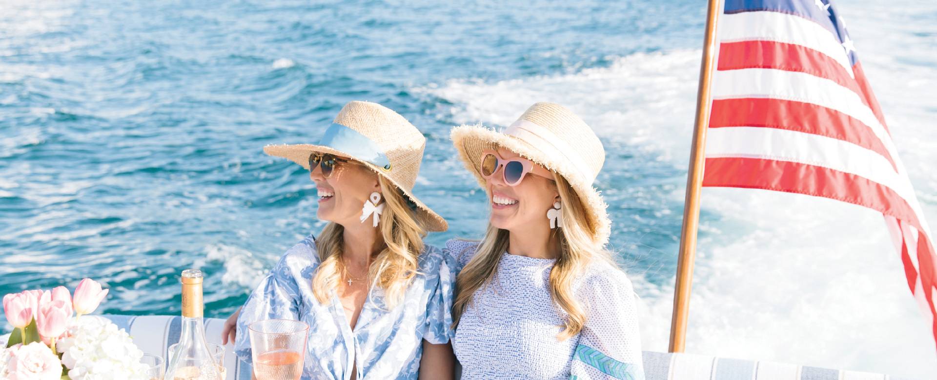 Two women enjoying a boat ride on open water, wearing sun hats and smiling as an American flag waves behind them.