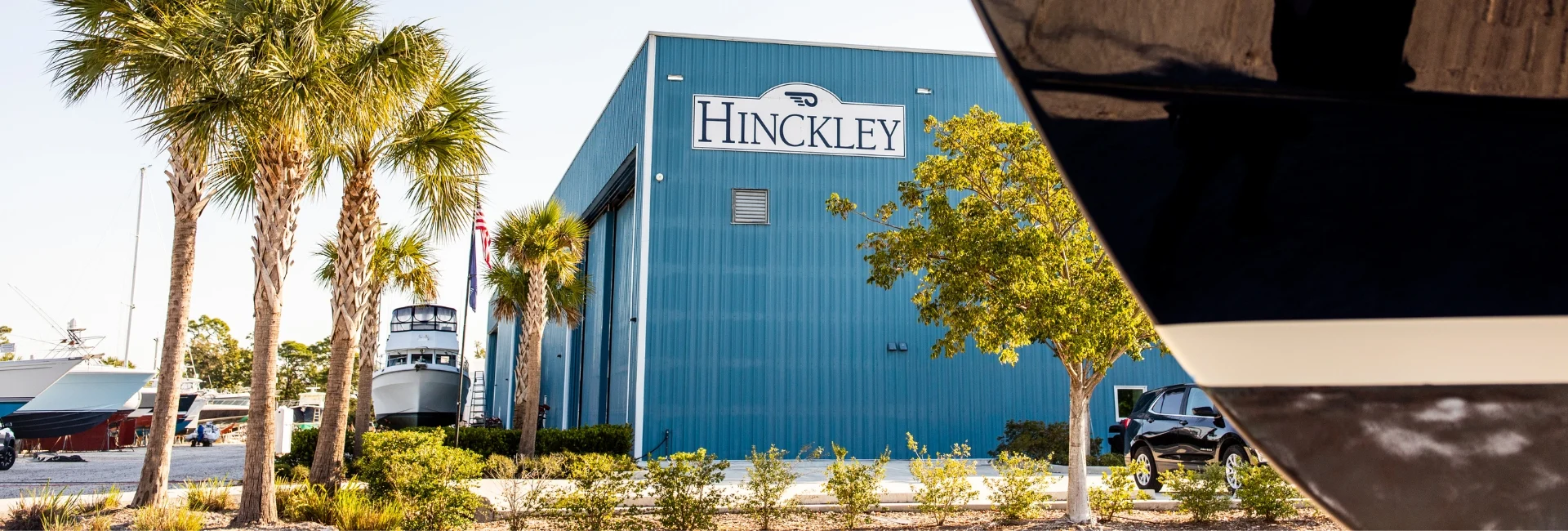 Exterior view of a Hinckley Yacht Services facility with palm trees, featuring the Hinckley name on the building and boats nearby.