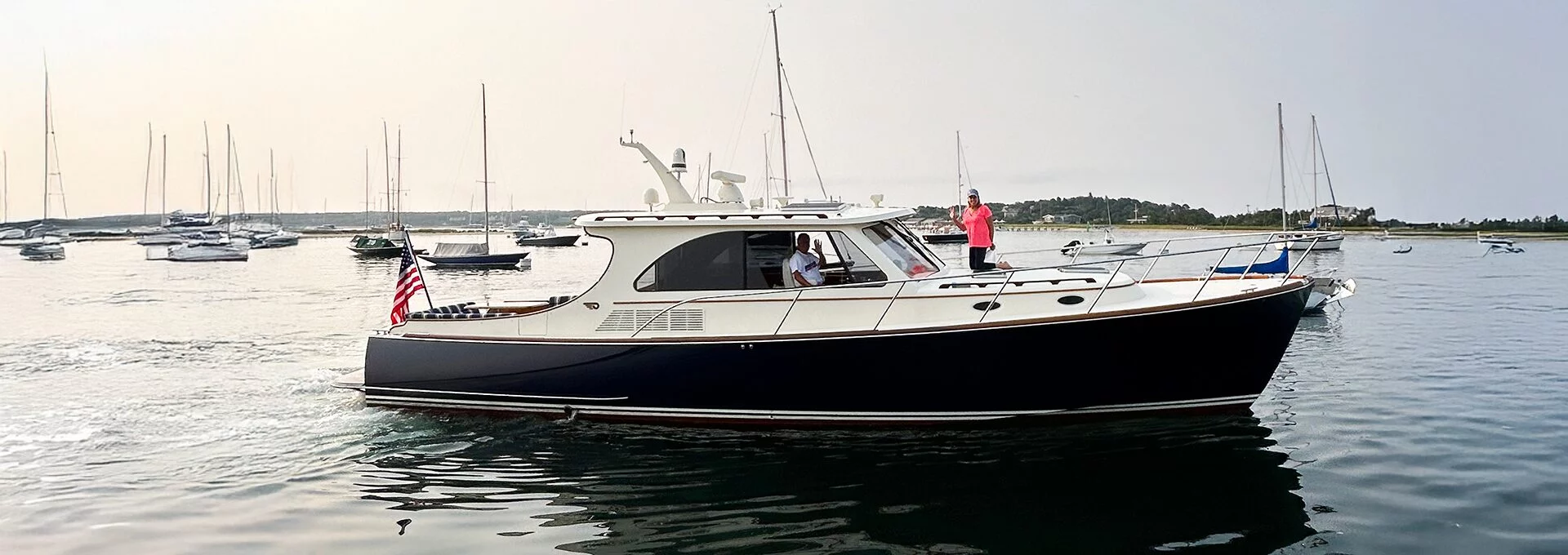 Hinckley Talaria 43 cruising calmly in a harbor, with people aboard and sailboats anchored in the background.
