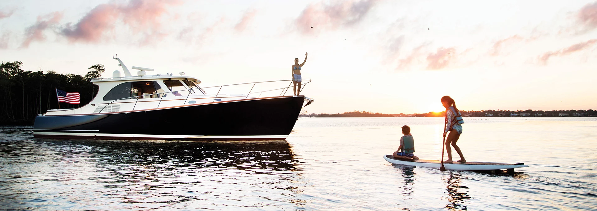Hinckley boat anchored on calm water at sunset while two people paddleboard nearby.
