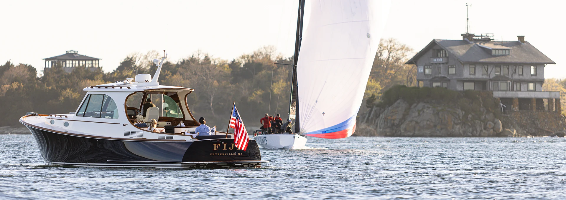 Hinckley motor yacht cruising on open water with a sailboat and shoreline building in the background.